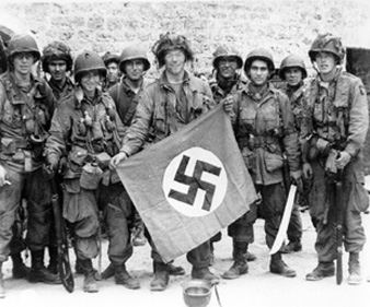 Photo:  SC189920 - American paratrooper, among the first to make successful landings on the continent, holds a Nazi flag captured in a village assault. Utah Beach, St. Marcouf, France. 8 June 1944