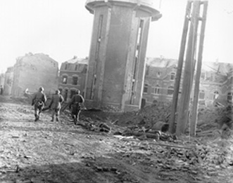 Photo:  SC200476 - Members of the 101st Airborne Division walk past dead comrades, killed during the Christmas Eve bombing of Bastogne, Belgium, the town in which this division was besieged for ten days. This photo was taken on Christmas Day. 1944